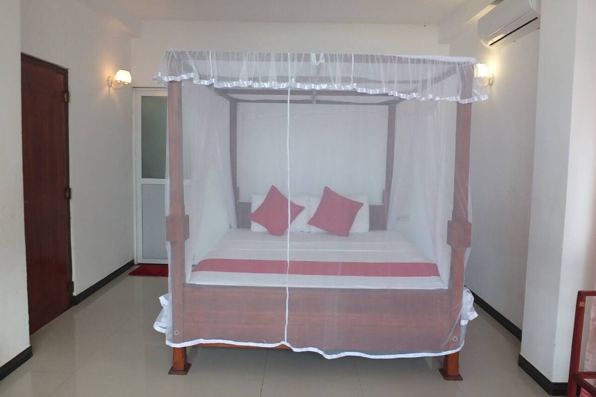 Uma cama com dossel, duas almofadas e um cobreleiro vermelho em um quarto com paredes brancas e uma porta de madeira.