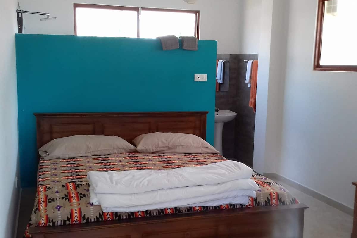 A bedroom with a wooden bed, teal walls, a window, and a bathroom visible through an open door.