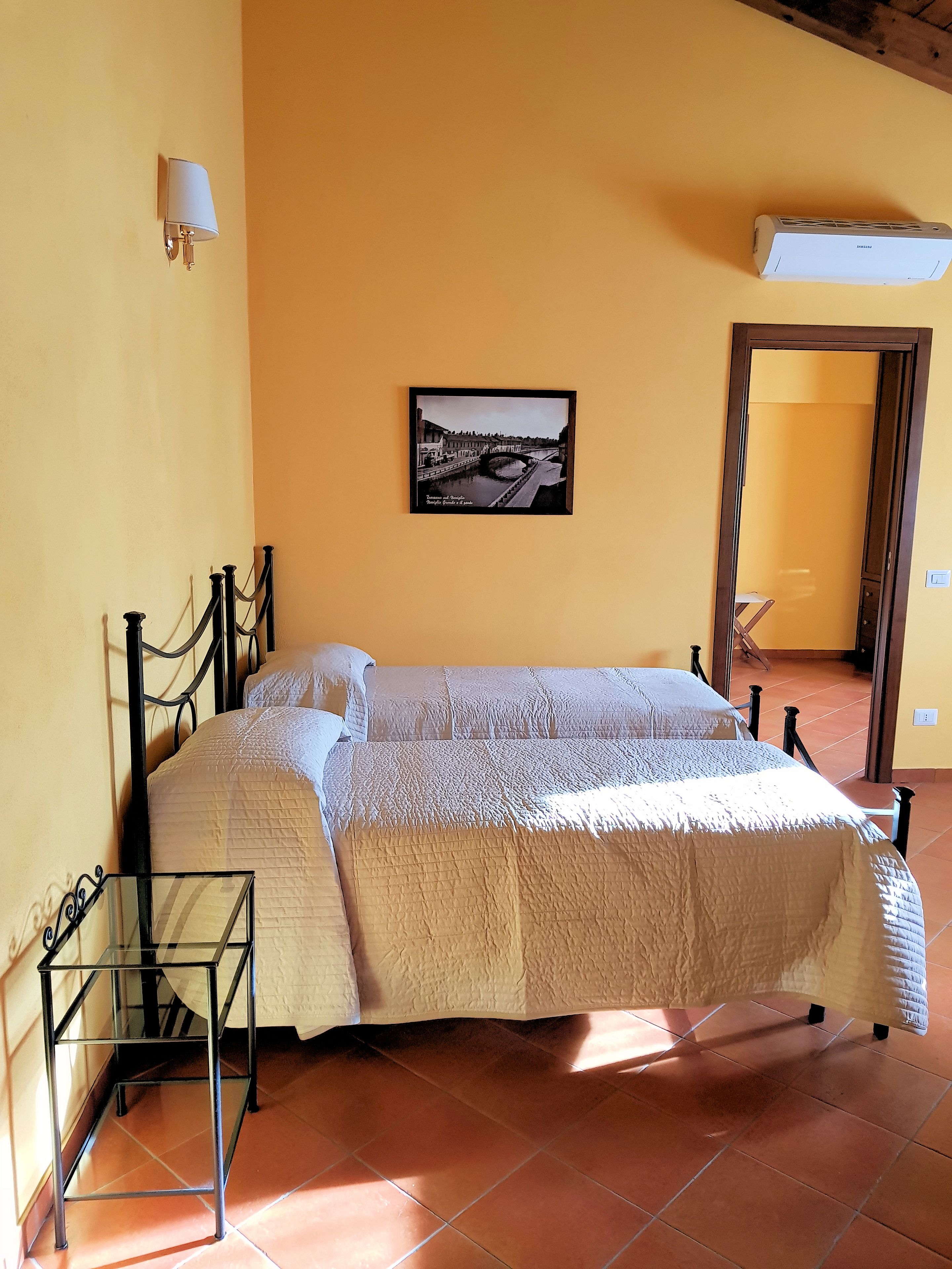 Quarto com cama, mesa de cabeceira, luminária na parede, quadro emoldurado e aparelho de ar condicionado.