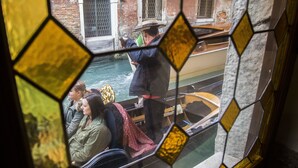 Une vue à travers un vitrail représentant un canal avec des bateaux, une personne vêtue d’un costume traditionnel et un bâtiment doté de fenêtres en arc de cercle.