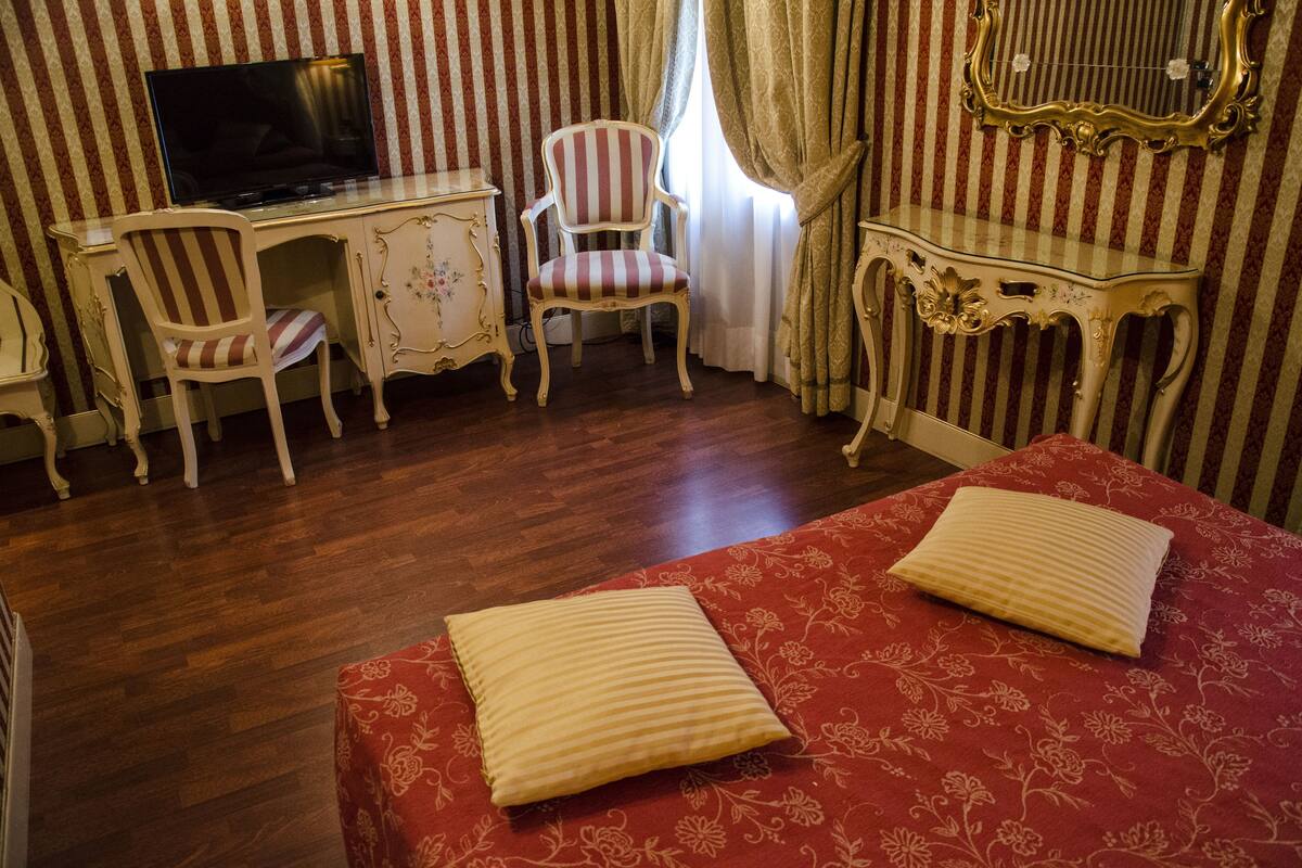 Habitación de hotel con piso de madera, un televisor sobre un escritorio, un sillón a rayas, una cama con colcha floral roja y un espejo en la pared.