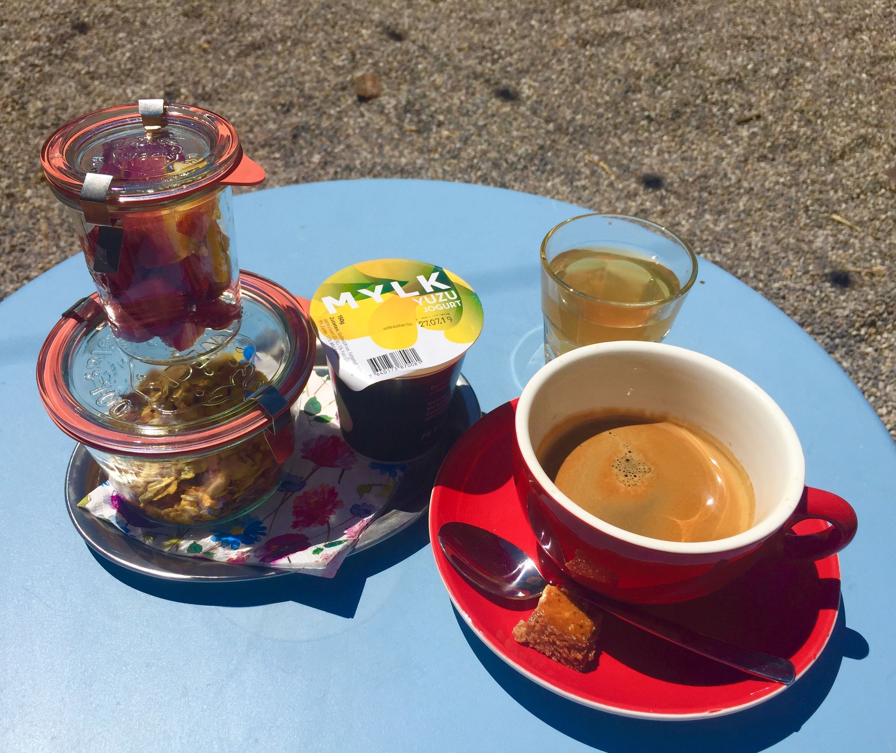 A table with a cup of coffee, a glass of tea, a jar of jam, and a container of yogurt.