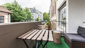 A balcony with a wooden table and bench, a wicker sofa, and a view of buildings and trees. - Hyve Hostel Basel (Basel)