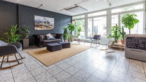 A modern lounge area with a dark brick wall, a large painting, and a seating area with a sofa and ottoman. There are several potted plants and a chalkboard menu visible. - Hyve Hostel Basel (Basel)