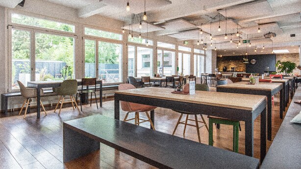 A spacious restaurant with high ceilings, exposed ductwork, and pendant lighting. Large windows provide natural light and views of the outdoors. - Hyve Hostel Basel (Basel)
