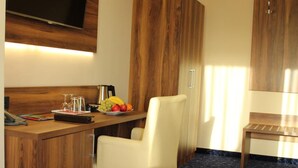 A hotel room with a wooden desk, a flat-screen TV mounted on the wall, a dining table with chairs, and a dark patterned carpet. - Grand Hotel Empire Frankfurt (Frankfurt)