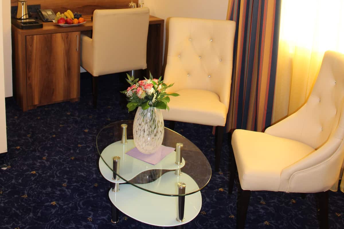 A hotel room with a flat-screen TV mounted on the wall, a dining area with a fruit bowl, a glass coffee table with a vase, two white armchairs, and a striped curtain.