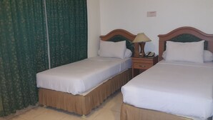 A hotel room with two single beds, a nightstand with a lamp, and a green curtain. - Hotel Panakkukang (Makassar)