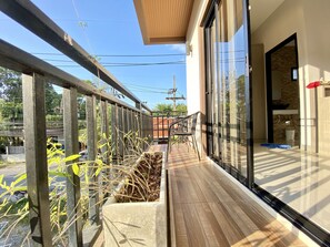 Uma varanda com piso de madeira, guarda-corpo de metal e vista para a rua e árvores.