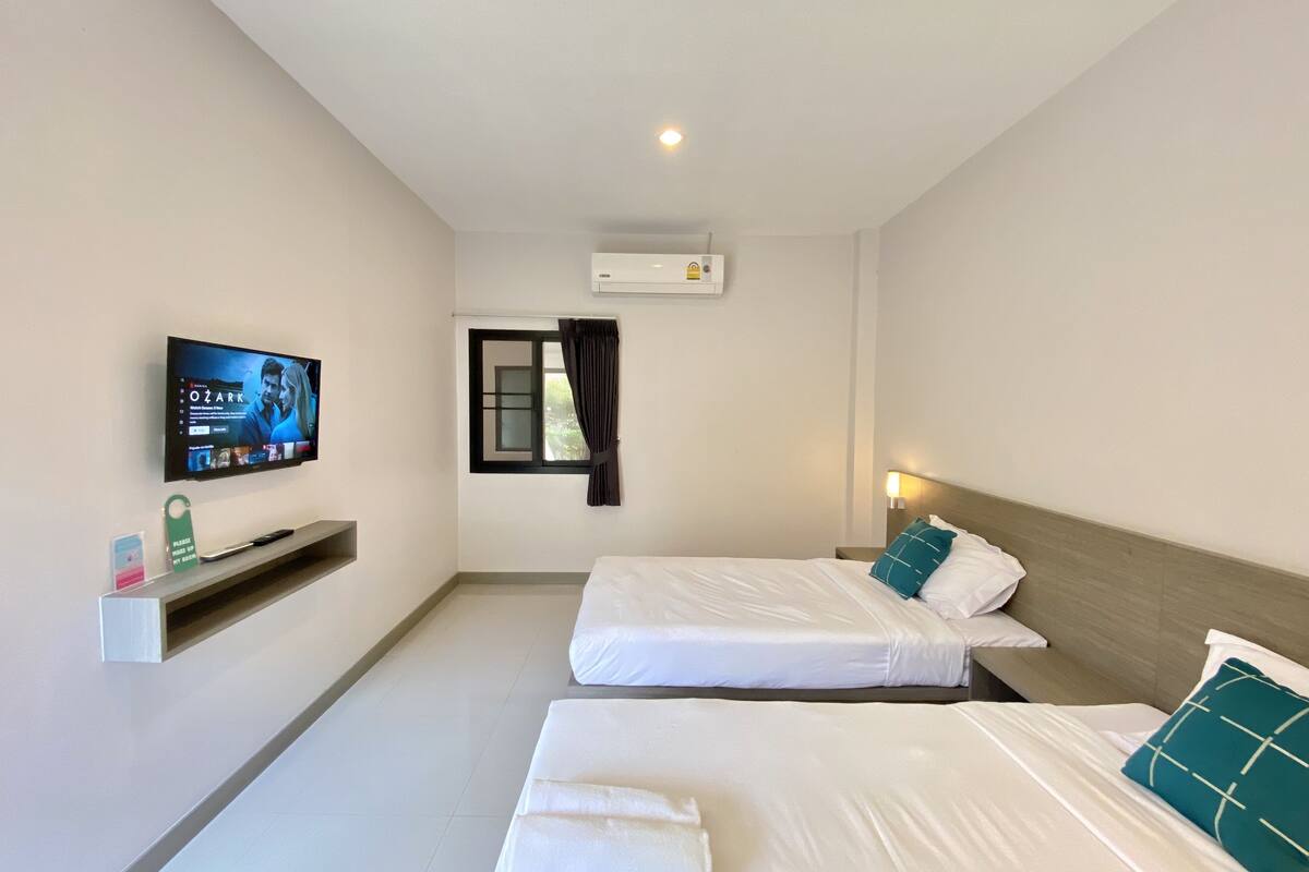 A hotel room with two single beds, a television mounted on the wall, a window with curtains, and a small shelf with a remote control.