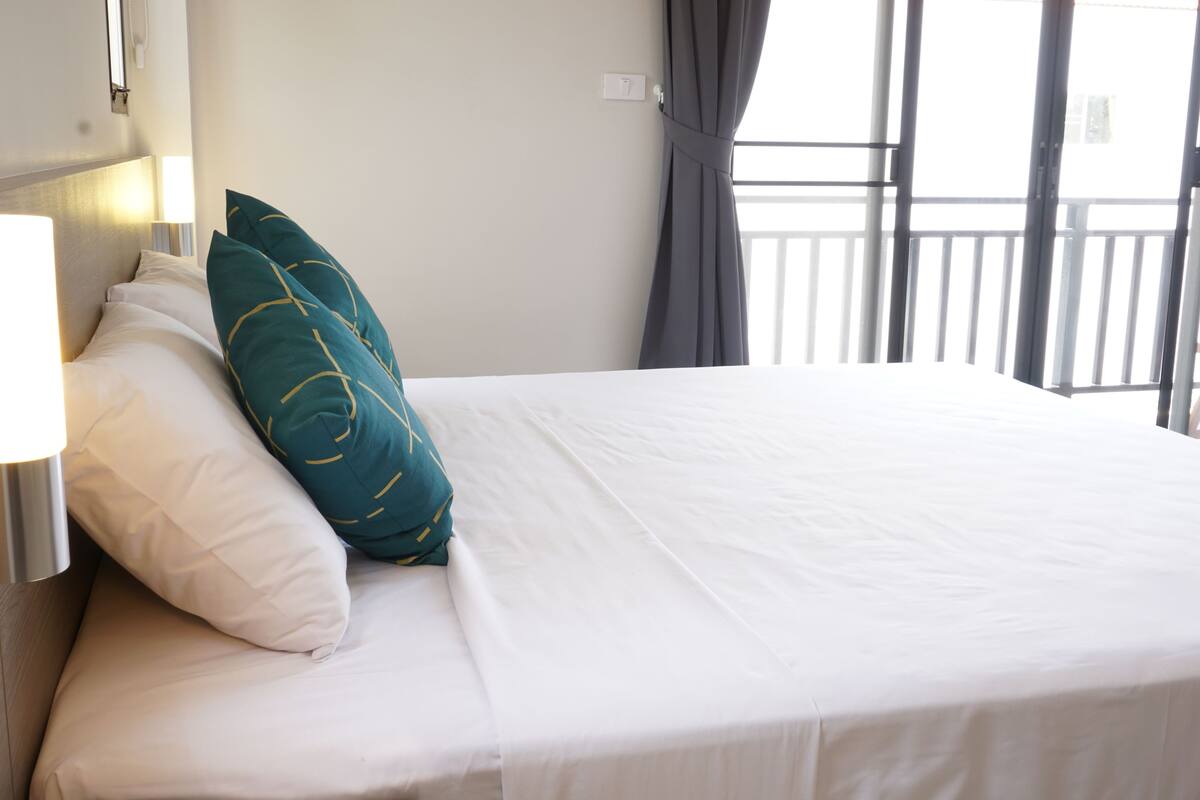 A hotel room with a neatly made bed, a bedside lamp, and a view of the balcony through the window.