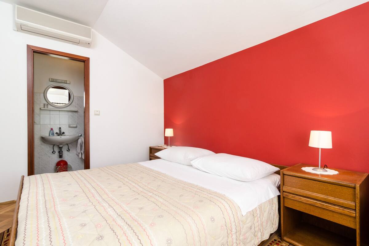 A bedroom with a bed, bedside tables, a nightstand, a red wall, a white ceiling, and a bathroom with a sink and mirror.