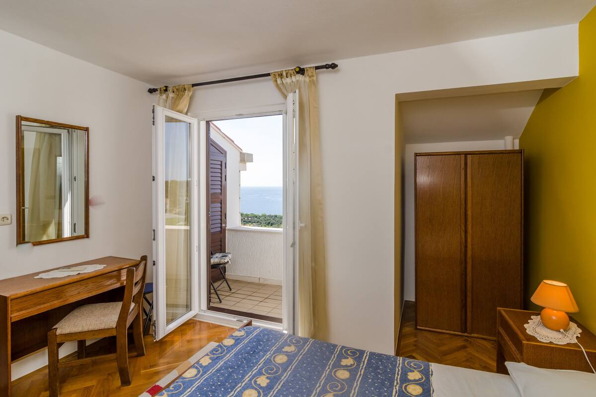A bedroom with a wooden desk, a chair, a bed with a patterned bedspread, a nightstand with a lamp, a wardrobe, and a balcony with a view of the sea.