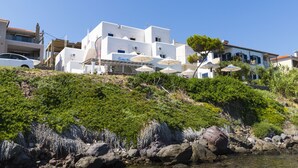 Un bâtiment blanc doté de nombreux balcons et agrémenté de parasols, situé sur une colline au terrain rocheux et à la végétation luxuriante.