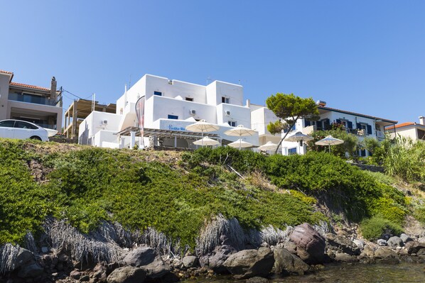 A white building with multiple balconies and outdoor umbrellas, situated on a hillside with rocky terrain and greenery. - Perdika Mare Guesthouse (Aegina)
