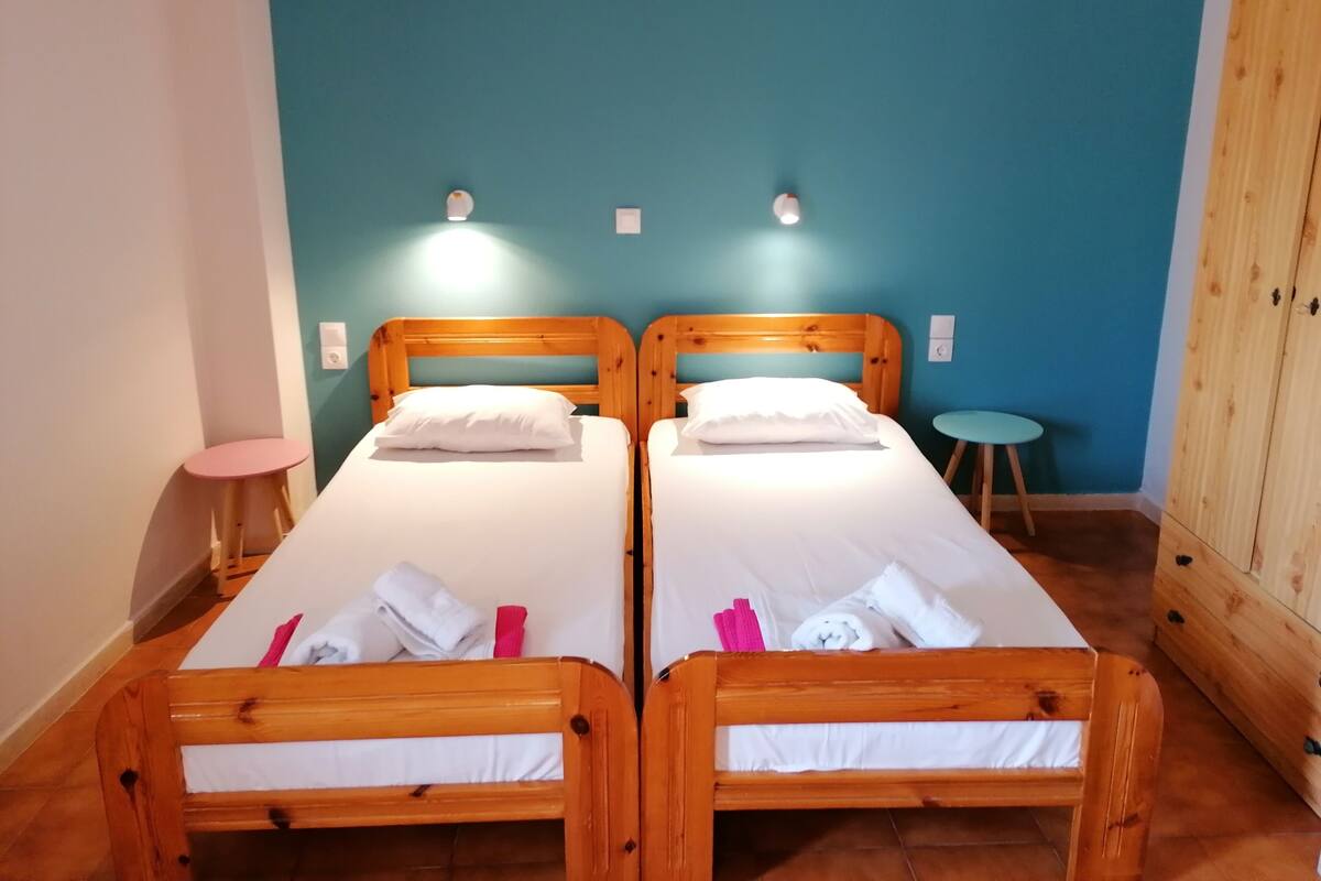 Twee houten bedden met witte lakens, een klein rond tafeltje en een kledingkast in een kamer met turquoise wanden.