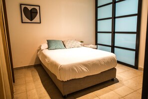 Ein ordentlich bezogenes Bett mit weißer Decke und zwei Kissen, einer herzförmigen Wanddekoration und einem großen Fenster mit Blick.