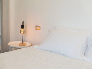 A bed with white linens, a round bedside table with a lamp, and a wall switch.