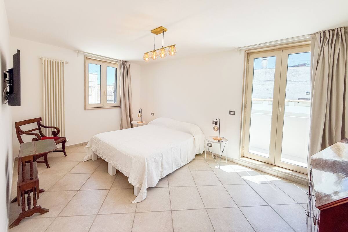Un dormitorio con cama, una silla, un escritorio, una lámpara y una ventana con cortinas.