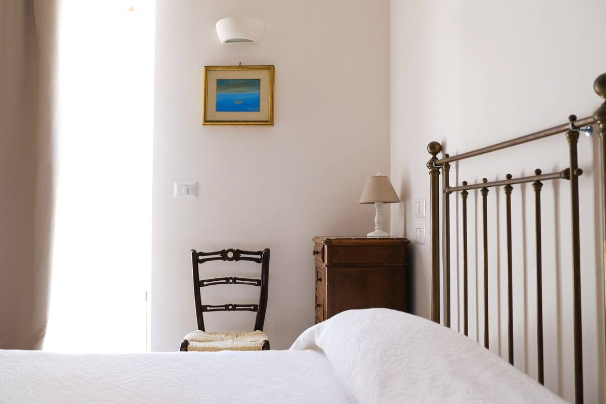 Un dormitorio con cama, una silla, una mesita de noche, una lámpara y un cuadro enmarcado en la pared.