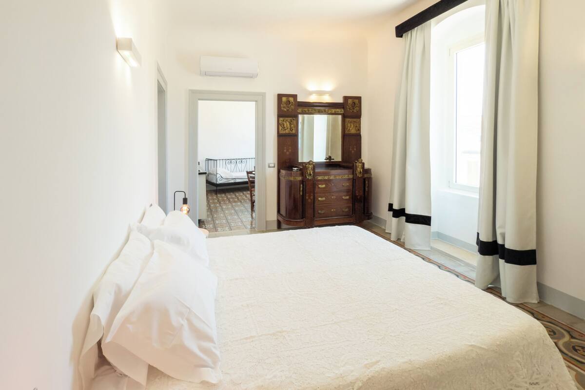 Una habitación de hotel con una cama grande, un aparador de madera, un espejo y una ventana con cortinas.