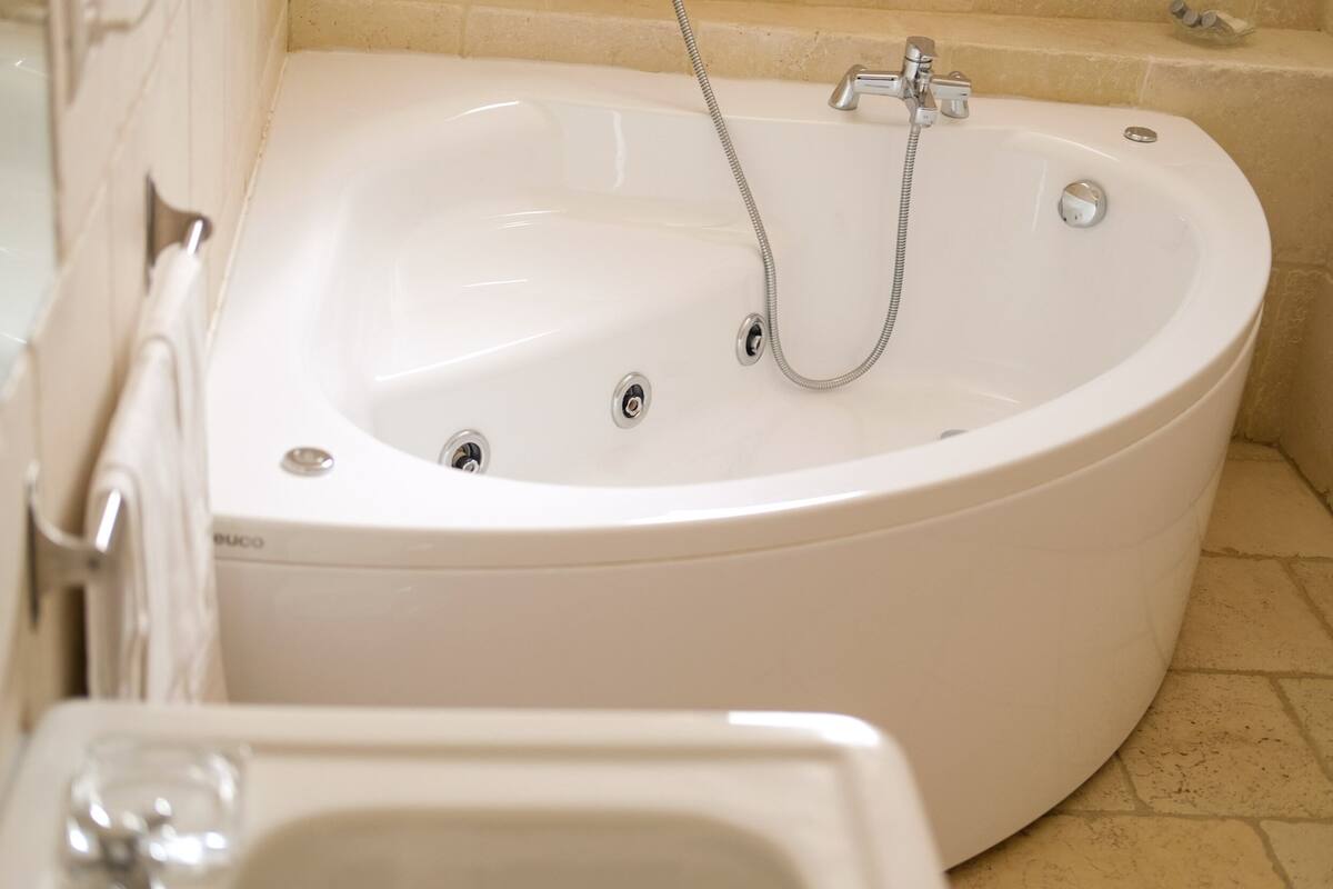Una bañera blanca con cabezal de ducha y grifos, un inodoro y un lavamanos en un baño con baldosas color crema.