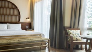 A hotel room with a large bed, a wooden headboard, two bedside lamps, a small table with a chair, and a view of the city through large windows. - Tints of Blue Hotel (Bangkok)