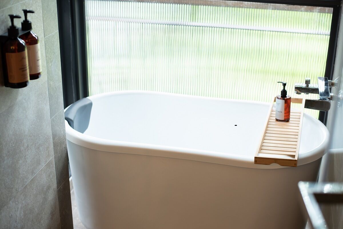 A modern bathroom with a freestanding bathtub, a wooden shelf with toiletries, and a view of a green landscape through a window.