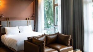 A hotel room with a bed, a leather sofa, a wooden coffee table, and a large window with curtains. - Tints of Blue Hotel (Bangkok)