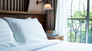 A neatly made bed with white linens, a wooden headboard, a bedside table with books, and a window with a view of greenery and buildings. - Tints of Blue Hotel (Bangkok)