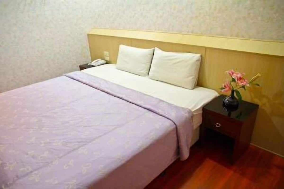 A neatly made bed with a light-colored headboard, a bedside table with a vase of flowers, and a wooden floor.