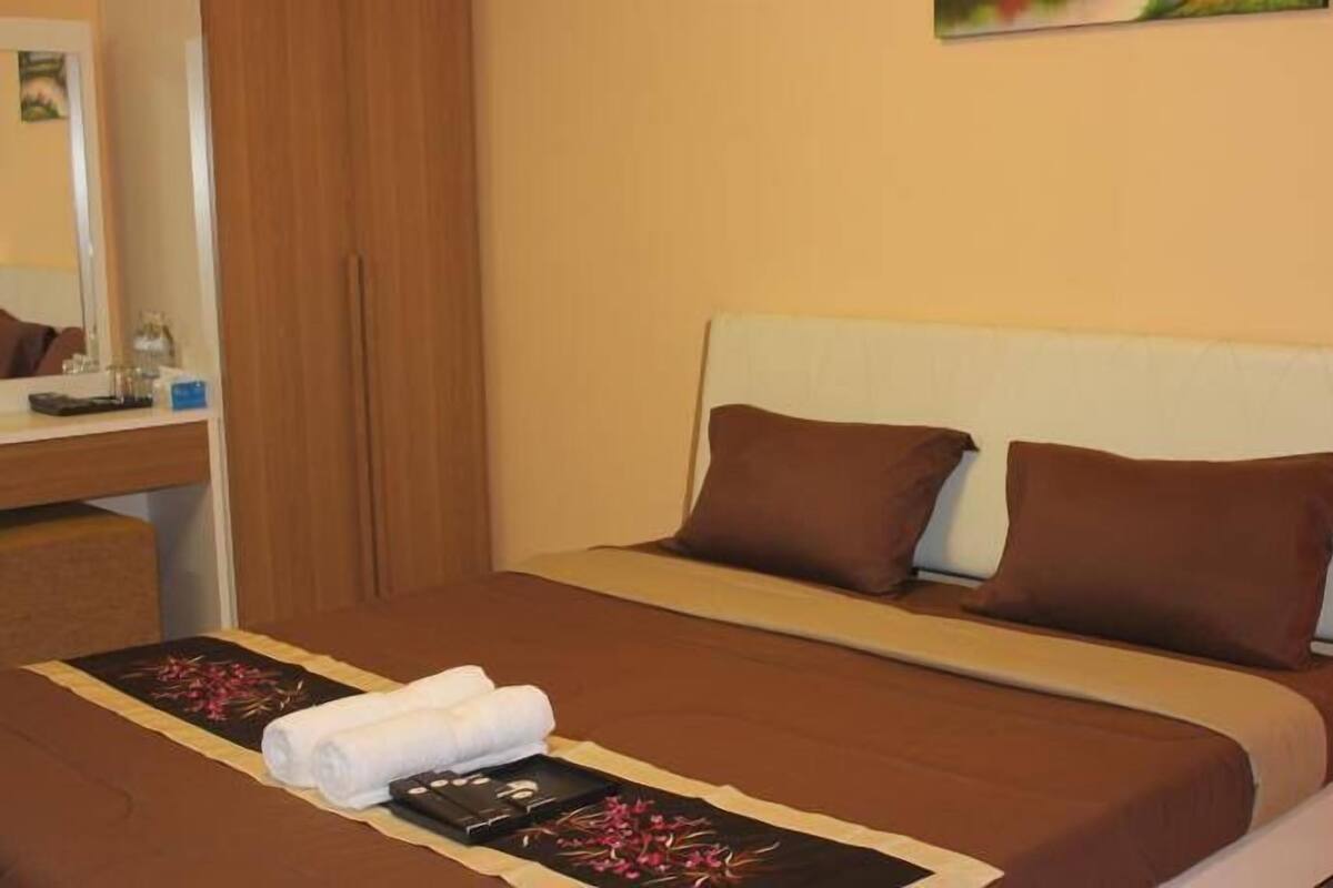 A neatly made bed with a brown bedspread and two pillows, a bedside table with a lamp, and a bathroom visible in the background.