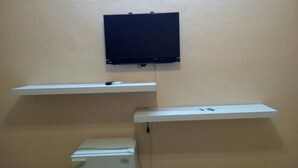 Una habitación con un refrigerador blanco, dos estantes blancos y un televisor negro montado en la pared.