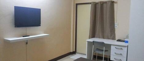 Una habitación pequeña y limpia, con una televisión de pantalla plana en la pared, un escritorio con una silla y una puerta corredera con cortina.