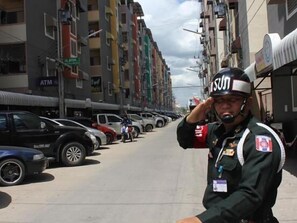 A street scene with a person in a uniform saluting, parked cars, and multi-story buildings with various signs. - Navavilla (Khlong Luang)