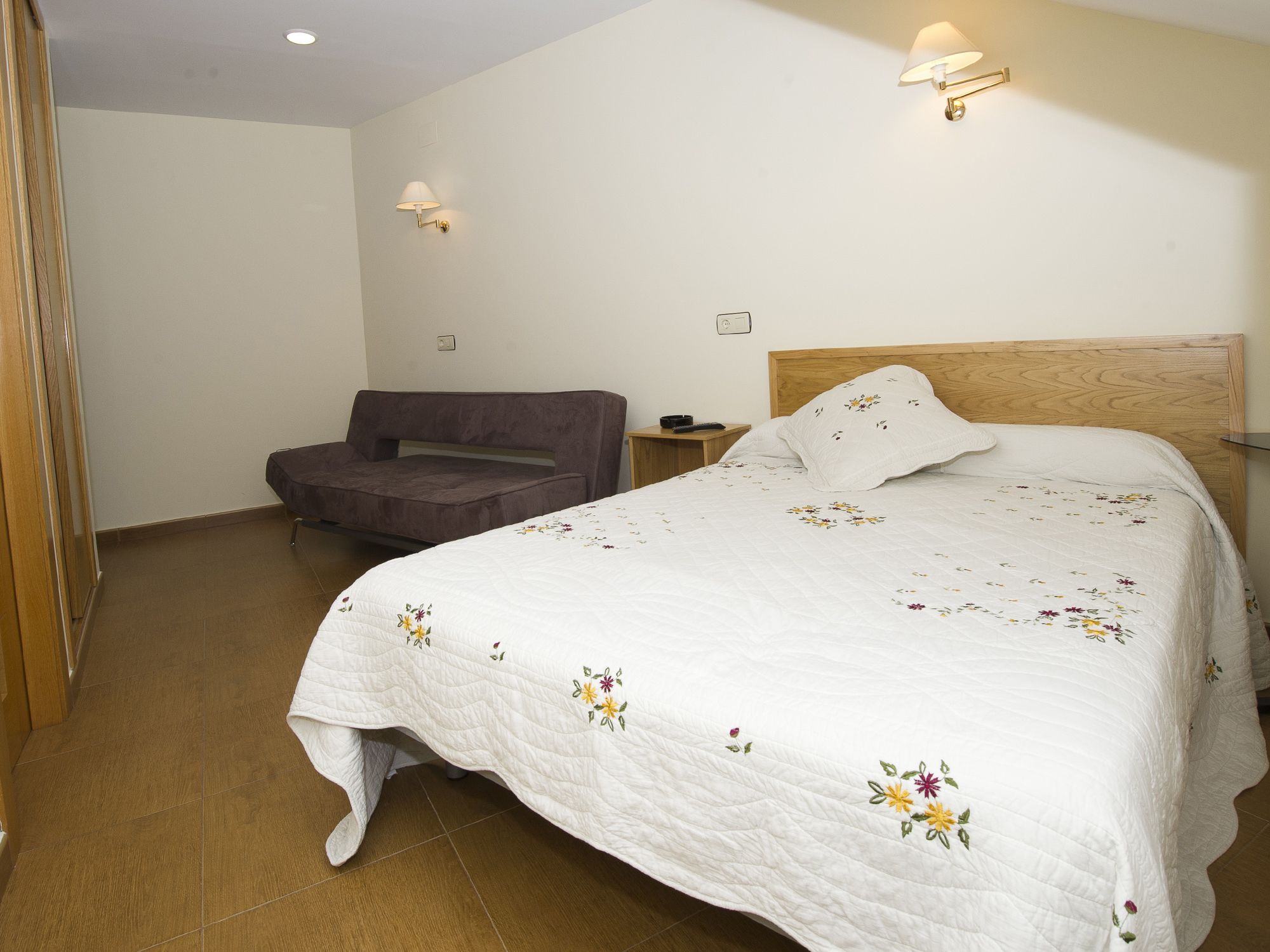 Un dormitorio con una cama grande, cabecera de madera, una mesita de noche, un sofá y dos lámparas de pared.