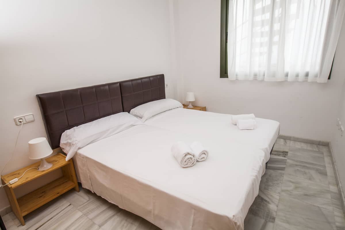 A neatly made bed with white linens and rolled towels, a bedside table with a lamp, and a power outlet on the wall.