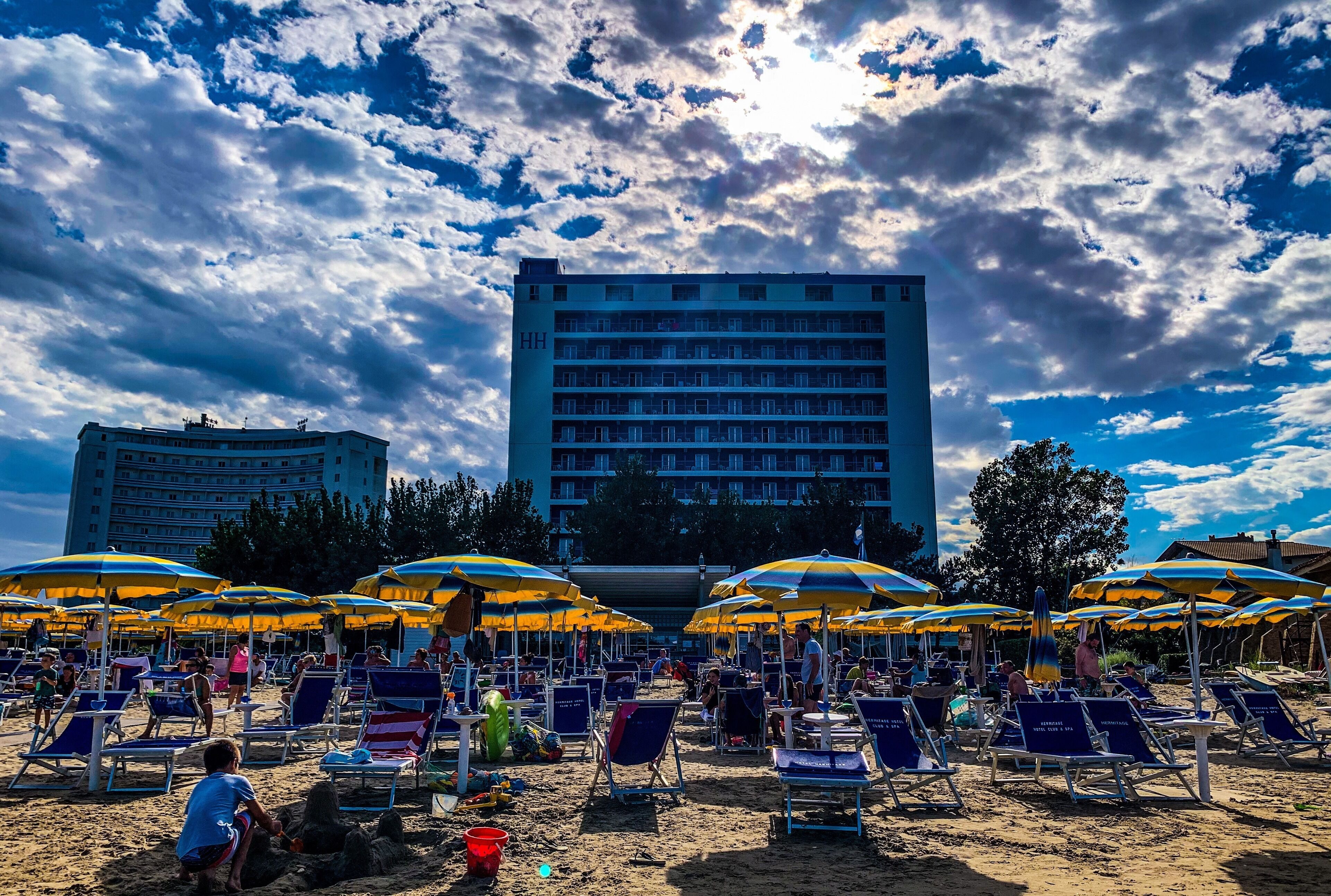 Une plage avec de nombreux parasols et des chaises longues, un grand bâtiment d’hôtel en arrière-plan et un ciel partiellement nuageux.