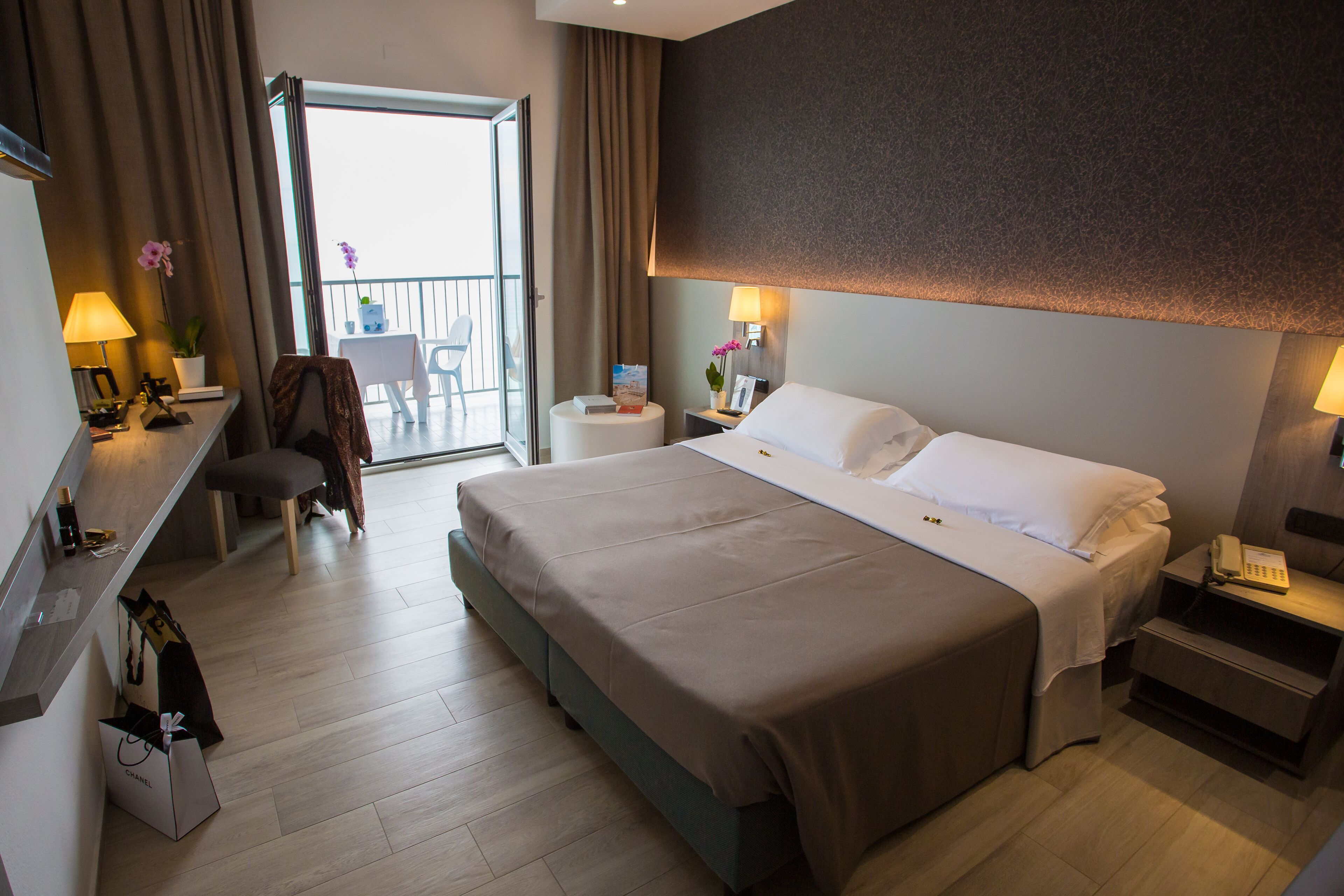 A hotel room with a large bed, a desk with a lamp, a chair, a small table with a plant, and a view of the balcony.
