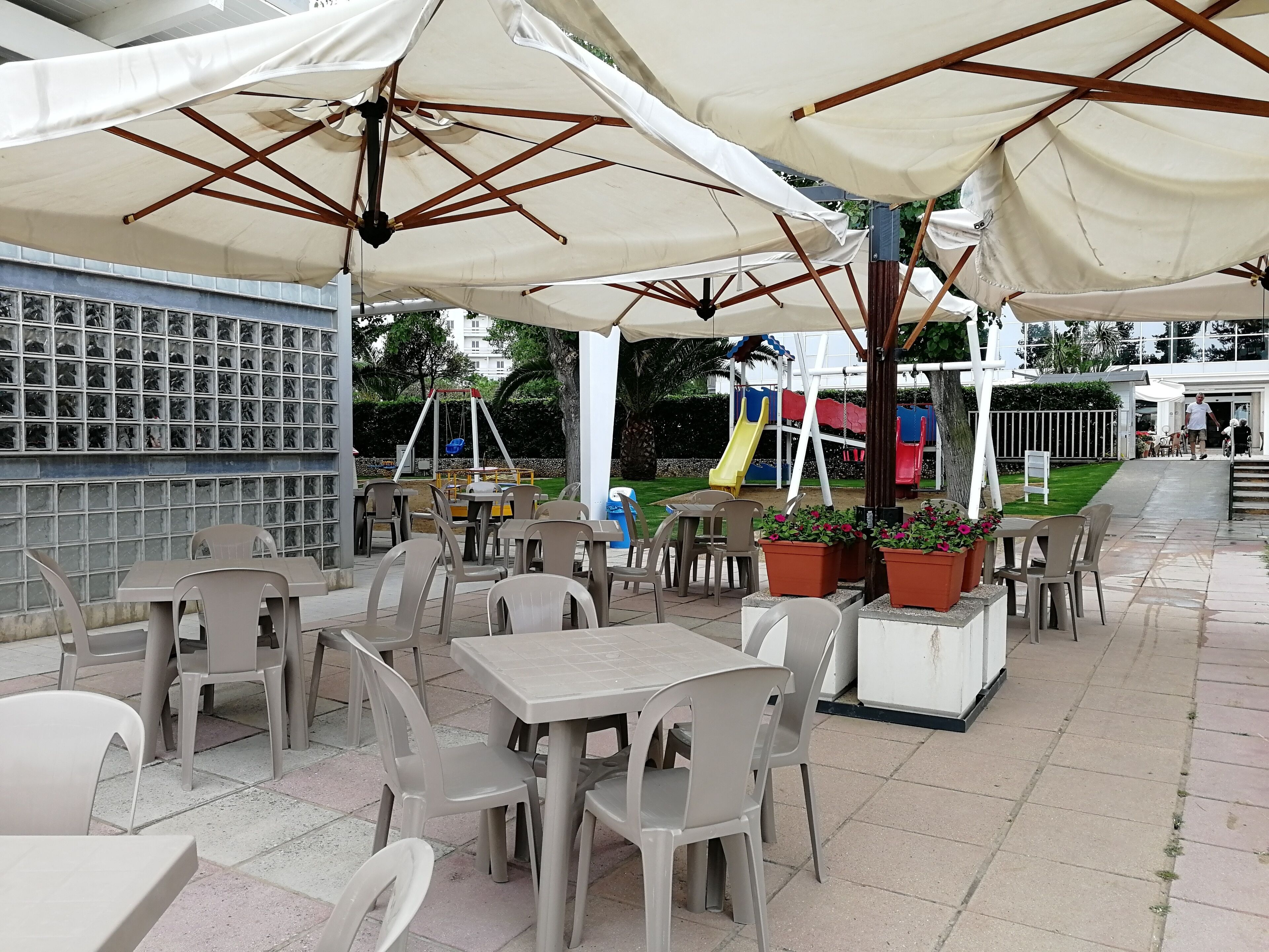 Area patio esterna con tavoli e sedie sotto grandi ombrelloni, una zona giochi per bambini sullo sfondo e una parete di vetro con motivo a griglia.