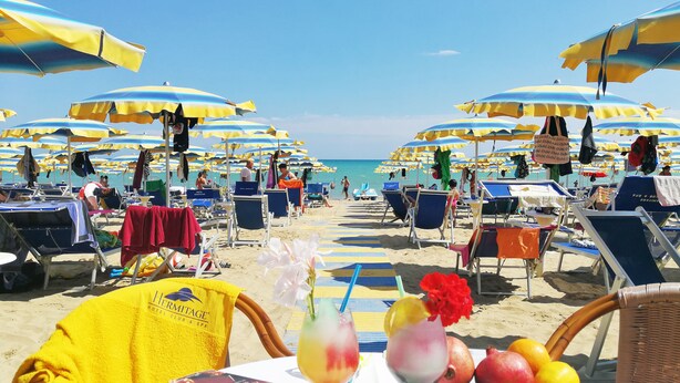 A beach with rows of sun umbrellas and lounge chairs, a table with a drink and fruit, and a view of the ocean. - Hermitage Hotel (Silvi)