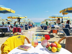 Een strand met rijen zonneschermen en ligbedden, een tafel met een drankje en fruit, en uitzicht op de oceaan.