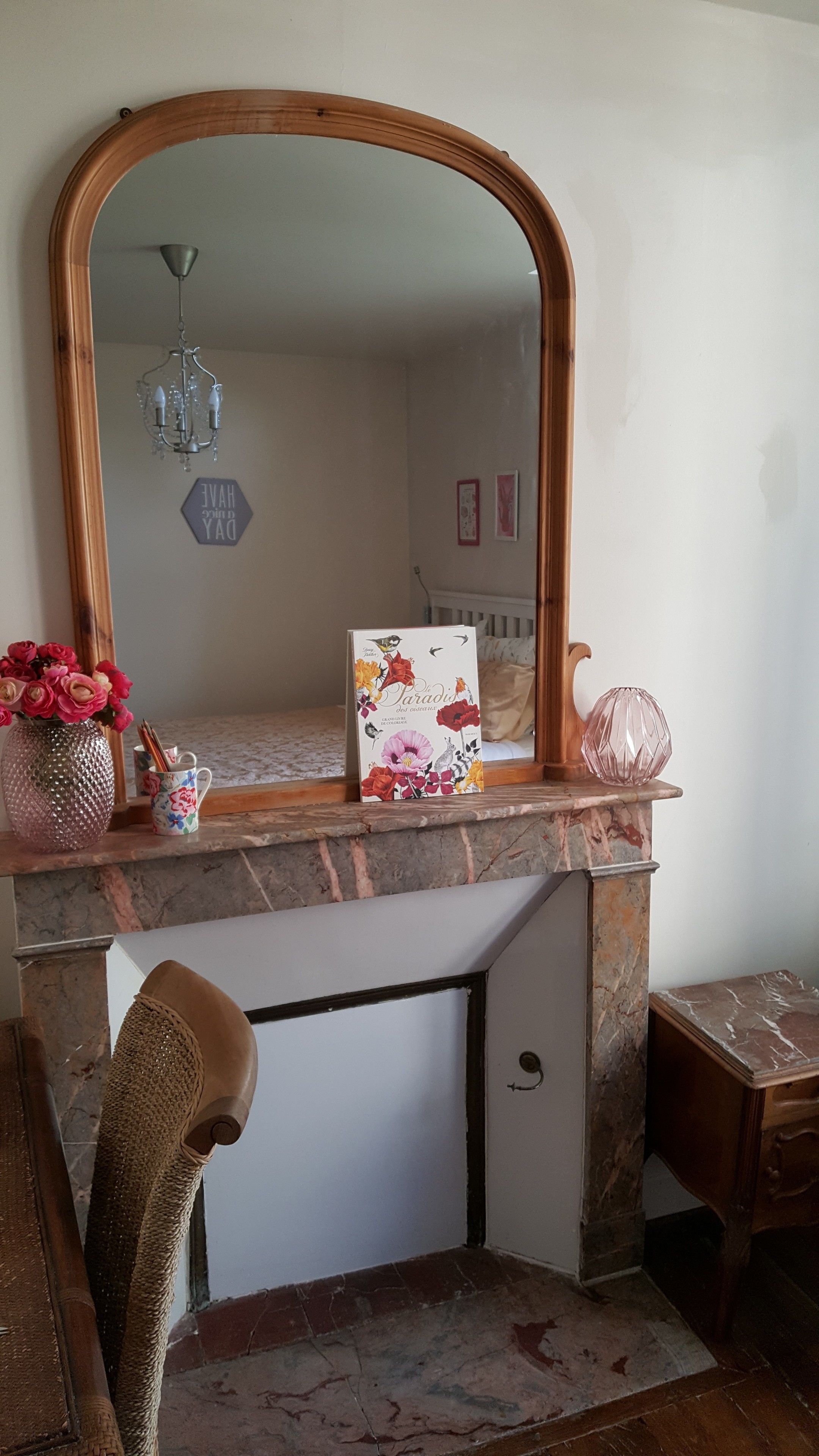 A room with a large mirror, a book titled 'Paradise', a vase with flowers, and a small table with a pink vase.