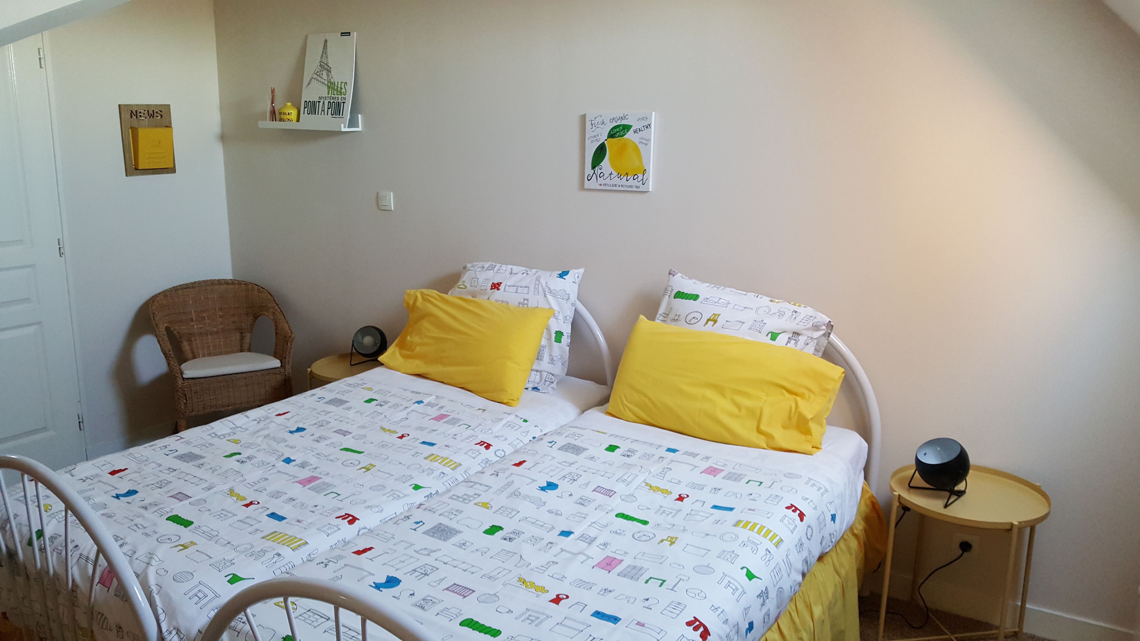 A bedroom with a bed, two yellow pillows, a wicker chair, a bedside table with a lamp, and a wall with a poster.