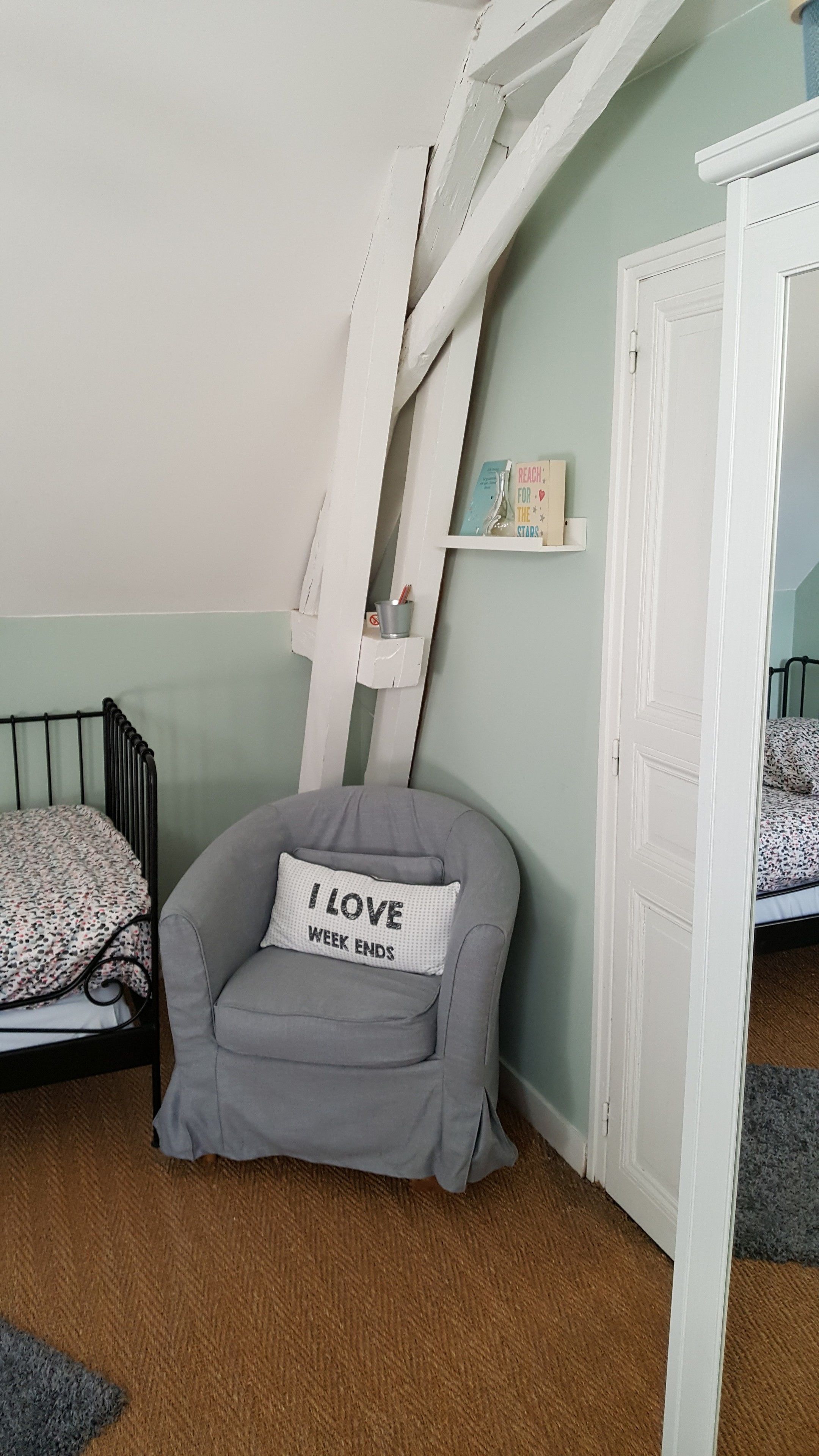 A cozy bedroom with a grey armchair, a small shelf with books, a white door, and a bed with patterned bedding.