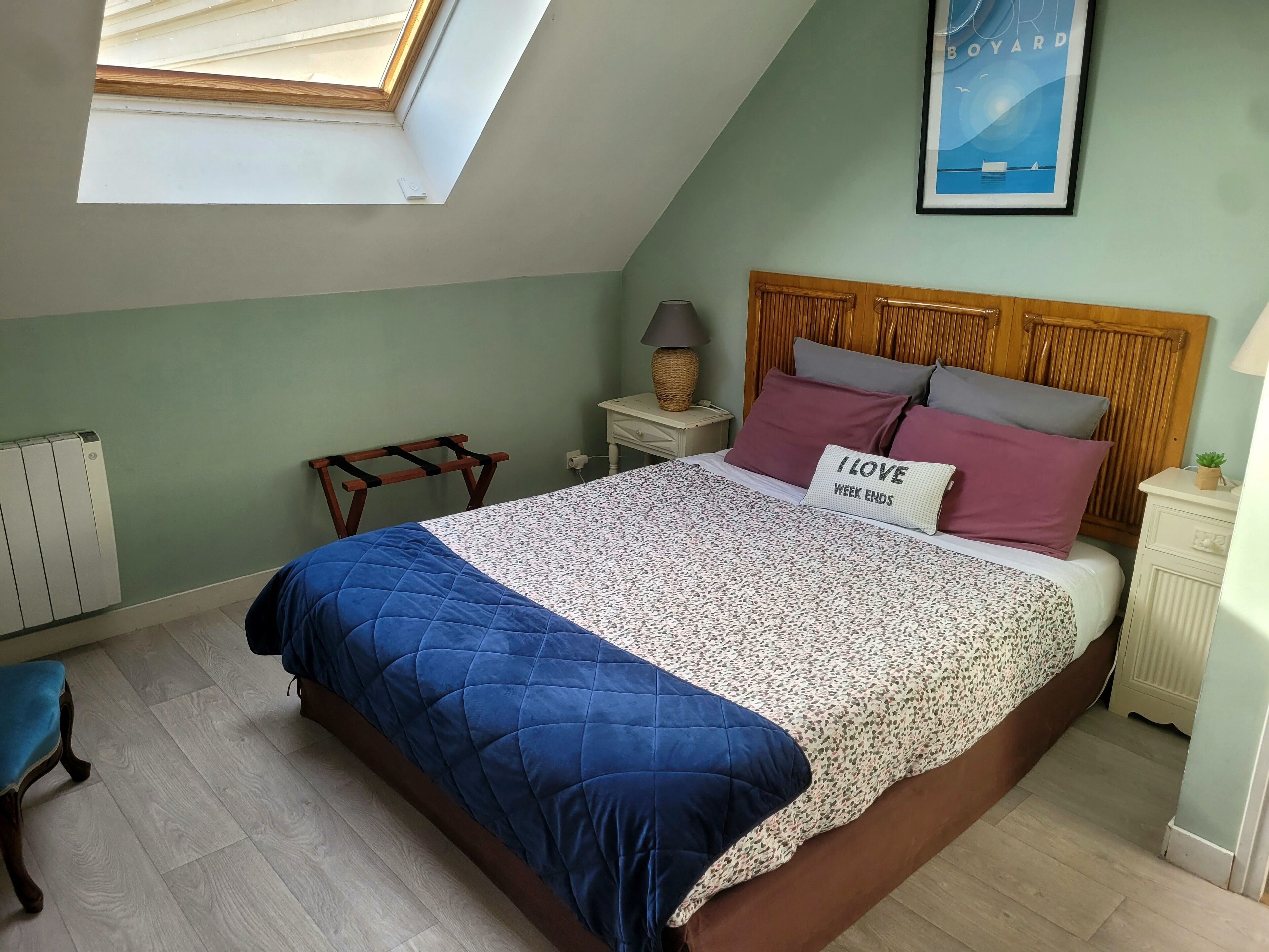 A bedroom with a sloped ceiling, a skylight, a bed with a blue quilt, a nightstand with a lamp, and a poster on the wall.