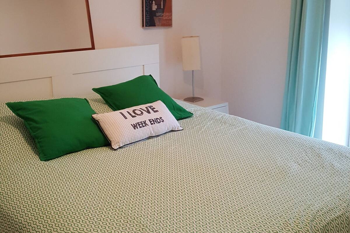 Een netjes opgemaakt bed met groen-wit beddengoed, een enkel groen kussen en een kussen met de tekst 'I LOVE WEEK ENDS'. Er is een witte nachtkastje met een lamp, een ingelijst beeld aan de muur en een hangende lamp.