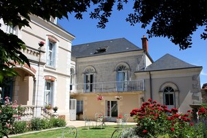 Um edifício de dois andares com varanda, um jardim com flores e uma mesa com cadeiras.