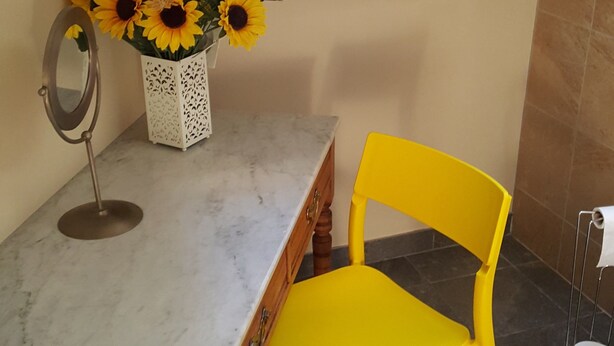 Ein Essbereich mit einem Marmortisch, einem gelben Stuhl, einer weißen Vase mit Sonnenblumen und einem auf einem Podest stehenden Spiegel.