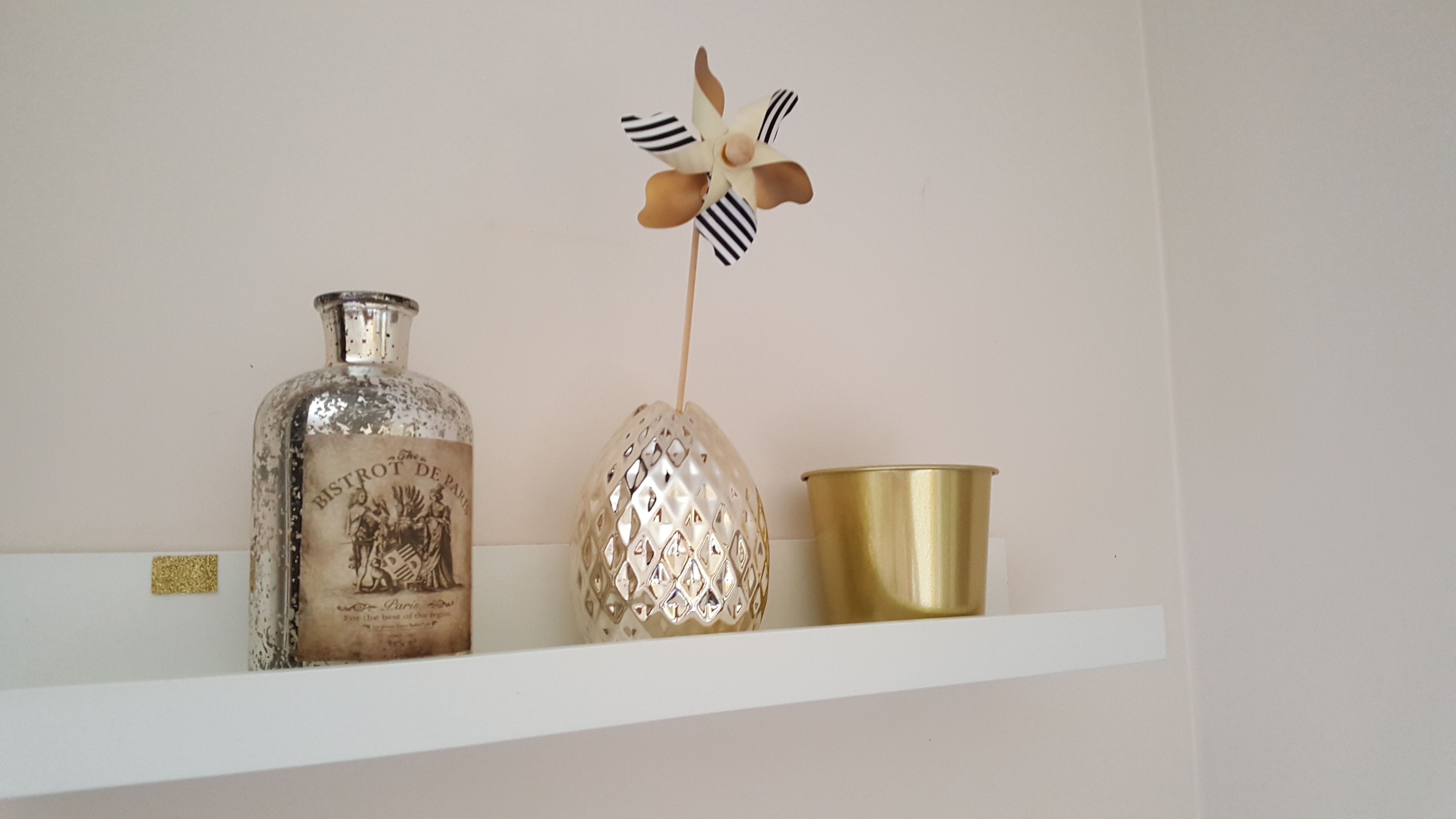 A shelf with a bottle, a decorative pinwheel, and a brass cup.